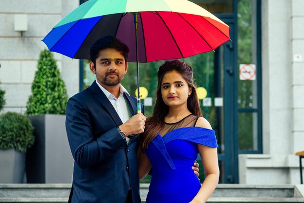 Mulher indiana em vestido azul longo à noite girando o tempo romântico junto com o namorado bonito amante sob o guarda-chuva multicolorido arco-íris europa urbana centro da cidade.