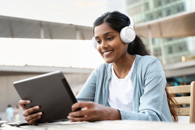 Foto mulher indiana sorridente usando fones de ouvido usando tablet digital assistindo vídeos sentado no café moderno