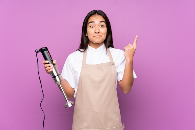 Mulher indiana usando o liquidificador na parede roxa, apontando com o dedo indicador uma ótima idéia