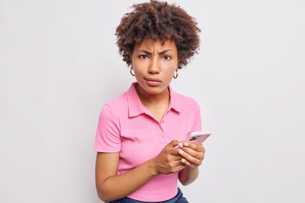 Mulher indignada e descontente com cabelo encaracolado usa telefone celular navega internet olha com raiva na frente usa camiseta rosa casual isolada na parede branca
