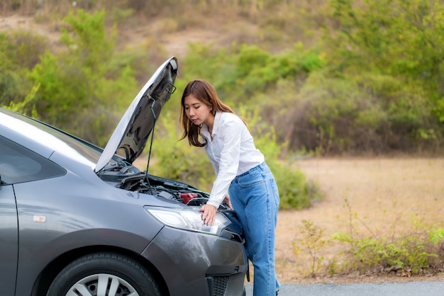 Mulher infeliz nova asiática que inspeciona o motor de automóveis quebrado na frente da capa aberta carro dividido no serviço de assistência rodoviária à espera da estrada secundária.