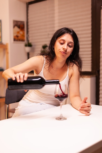 Mulher infeliz servindo vinho tinto da garrafa no vidro, sentado sozinho à mesa na cozinha. Pessoa infeliz, doença e ansiedade, sentindo-se exausta por ter problemas de alcoolismo.