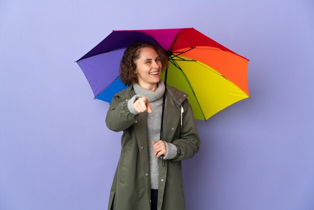 Mulher inglesa segurando um guarda-chuva isolado em um fundo roxo apontando para a frente com uma expressão feliz