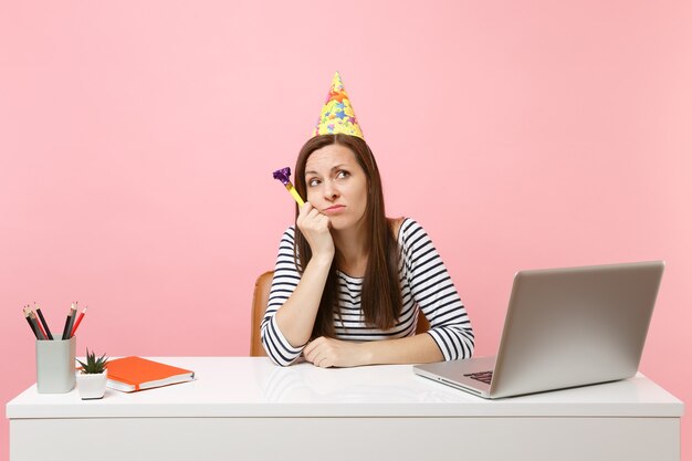 Mulher insatisfeita com chapéu de aniversário tocando cachimbo encostado na mão, ninguém veio comemorar, sente-se no trabalho na mesa branca com o laptop
