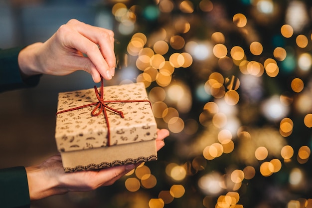 Mulher irreconhecível embrulha a caixa de presente sobre a árvore de Natal com luzes brilhantes e guirlandas Caixa de presente decorada nas mãos da mulher Conceito de férias e celebração