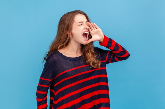 Foto mulher irritada vestindo suéter listrado estilo casual gritando alto, segurando a mão perto da boca, anunciando notícias importantes, atraindo a atenção. tiro de estúdio interior isolado sobre fundo azul.