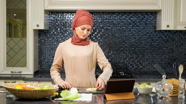 Mulher islâmica em hijab prepara jantar cortando legumes frescos assistindo receita de prato halal na internet usando laptop fechado