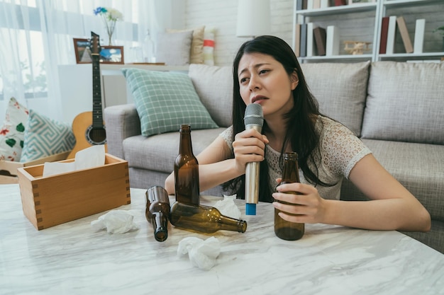 mulher japonesa asiática de coração partido, sentada ao lado de uma mesa com garrafas de cerveja e chumaços de ranho, está abusando de álcool enquanto chora cantando karaokê sozinha em casa após o término.