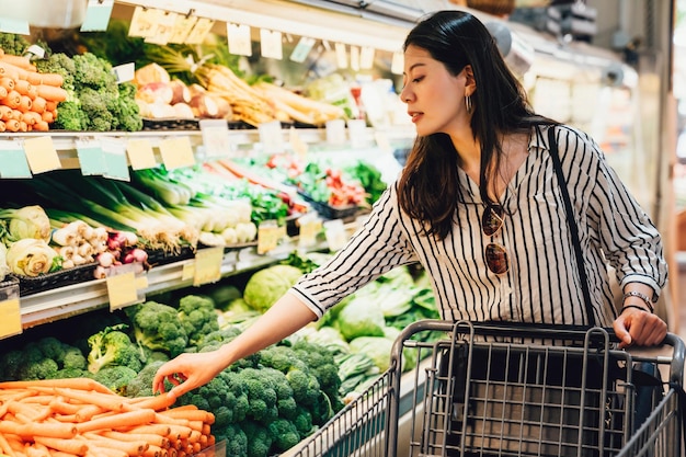 mulher japonesa no supermercado empurrando o carrinho de compras andando pela área de legumes e frutas. bela senhora asiática pegando cenouras ao lado de brócolis compra alimentos cuidados de saúde. esposa preparar para o jantar