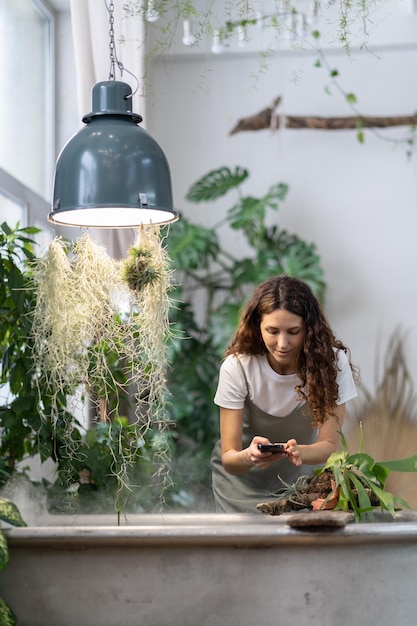 Mulher jardineira tirando foto de planta aquática em estufa usando smartphone sob banho autônomo