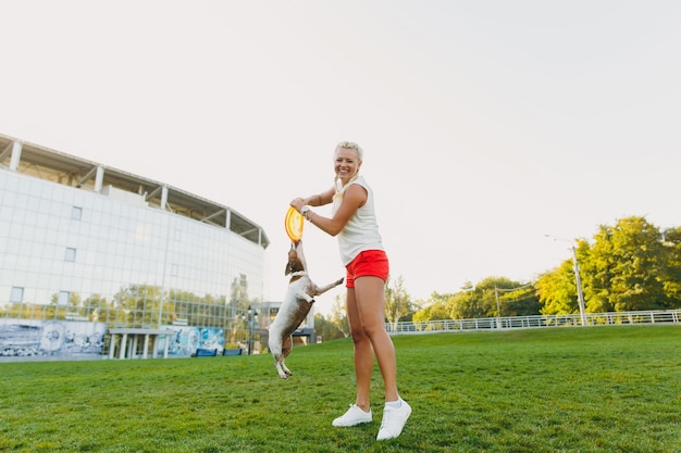 Mulher jogando o disco voador laranja para o pequeno cachorro engraçado, que pegá-lo na grama verde. pequeno animal de estimação jack russel terrier brincando ao ar livre no parque. cão e dono ao ar livre.