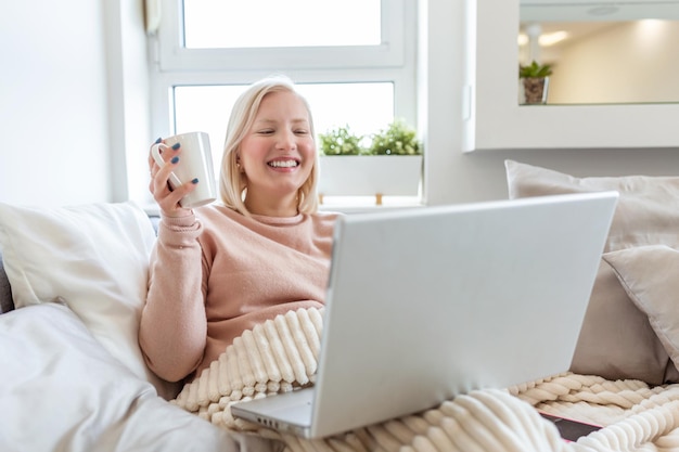 Mulher jovem a beber café em casa na cama e a verificar o seu portátil Mulher jovem e bonita a beber café na casa na cama enquanto trabalha no seu portátil a verificar o e-mail de manhã