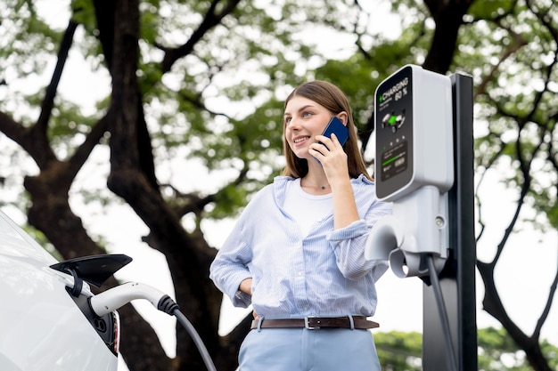 Foto mulher jovem a falar no smartphone enquanto recarga o carro elétrico exalt