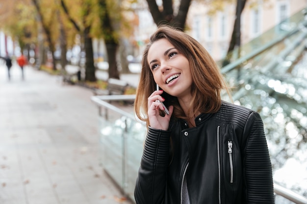 Mulher jovem adorável e feliz em pé e falando no celular na cidade