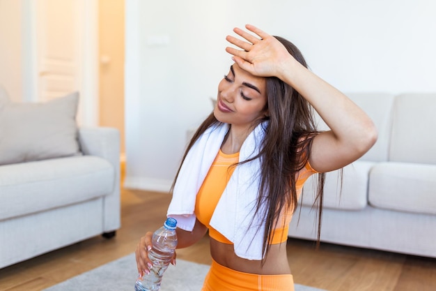 Mulher jovem adulta bebendo água de garrafa de plástico sentada em um tapete de fitness e descansando depois de treinar em casa Mulher jovem bonita limpando o suor com uma toalha exausta depois do treino diário