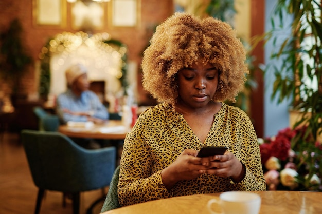Mulher jovem africana com penteado elegante usando telefone celular enquanto está sentado à mesa no café