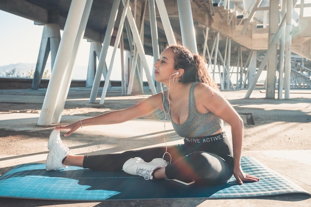 Mulher jovem africana poc de comprimento total, esticando a perna antes de iniciar o treinamento em um local de esporte urbano na cidade sobre um tapete de ioga. Novo hábito para o conceito de fitness de perder peso do ano