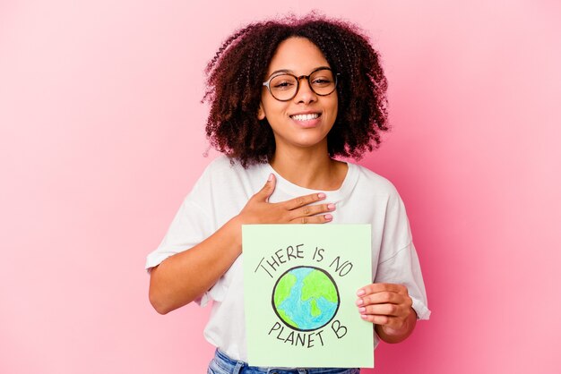 Mulher jovem afro-americana de raça mista segurando um papelão de conceito de proteção mundial ri alto, mantendo a mão no peito.