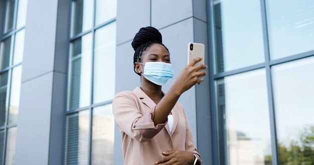 Mulher jovem afro-americana muito feliz na máscara médica tendo videochat no smartphone ao ar livre no edifício comercial. mulher bonita alegre falando e videochatting via webcam no celular.