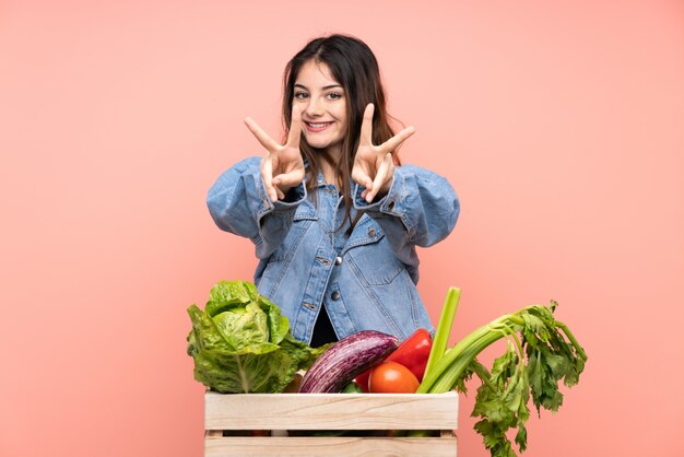 Mulher jovem agricultor segurando uma cesta cheia de legumes frescos, sorrindo e mostrando sinal de vitória