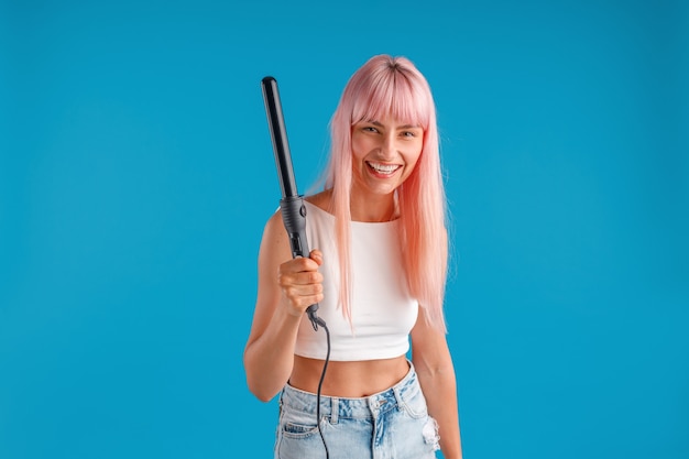 Mulher jovem alegre com cabelo rosa em uso casual sorrindo para a câmera segurando um modelador de cabelo em pé