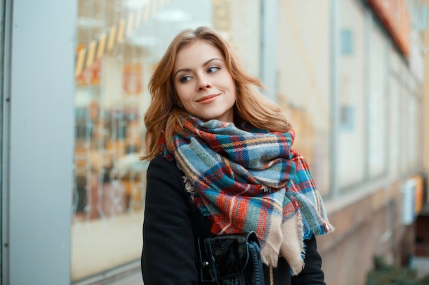 Mulher jovem alegre com um casaco preto quente de inverno com uma bolsa de couro e um lenço quente de lã na moda em uma gaiola perto de uma vitrine em uma cidade decorada com luzes festivas. menina feliz