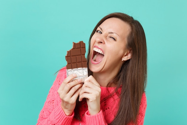 Mulher jovem alegre louca no suéter rosa de malha, segurando na mão e mordendo a barra de chocolate isolada no fundo da parede azul turquesa, retrato de estúdio. Conceito de estilo de vida de pessoas. Simule o espaço da cópia.