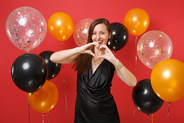 Mulher jovem alegre no vestido preto comemorando, mostrando o gesto do coração em balões de ar de fundo vermelho brilhante. São Valentim, dia da mulher, feliz ano novo, conceito de festa de feriado de maquete de aniversário.