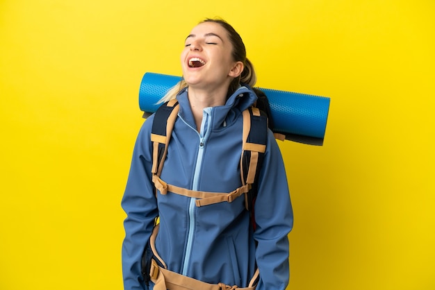 Mulher jovem alpinista com uma grande mochila sobre um fundo amarelo isolado rindo