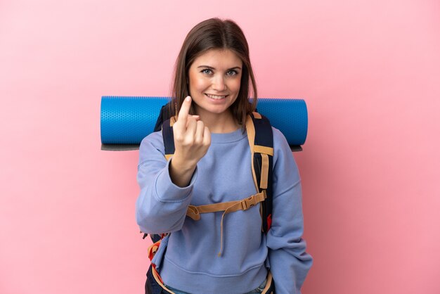 Mulher jovem alpinista com uma mochila grande isolada em um fundo rosa fazendo gesto de aproximação