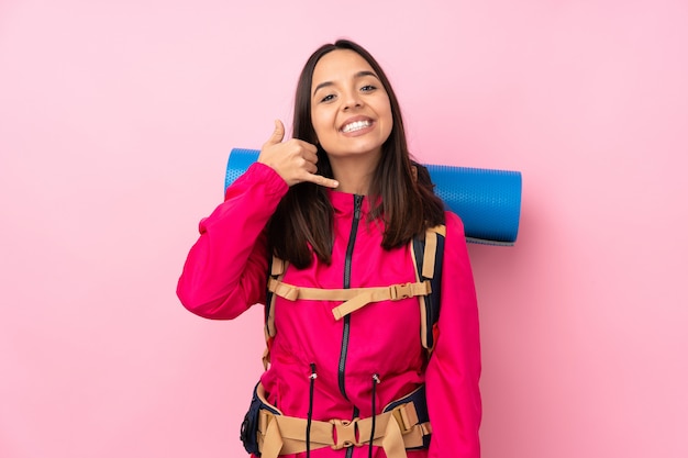 Mulher jovem alpinista com uma mochila grande sobre parede rosa isolada, fazendo gesto de telefone. Ligue para mim de volta