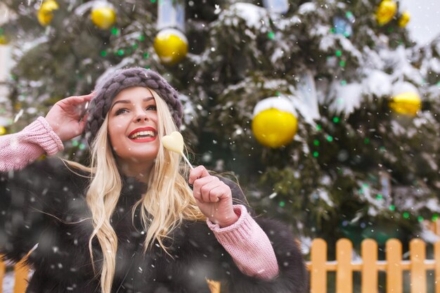 Mulher jovem animada com balas de chocolate caminhando perto da árvore do ano novo durante uma queda de neve