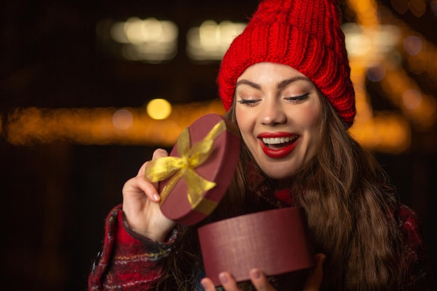 Mulher jovem animada com chapéu vermelho, abrindo a caixa de presente na feira de Natal à noite. Espaço vazio