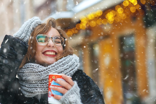 Mulher jovem animada de óculos com uma xícara de café, caminhando perto de vitrines decoradas em tempo de neve. Espaço vazio