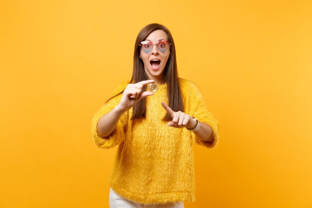 Mulher jovem animada em copos de coração apontando o dedo indicador em bitcoin, moeda de metal de cor dourada, futura moeda isolada em fundo amarelo. Pessoas sinceras emoções, estilo de vida. Área de publicidade.