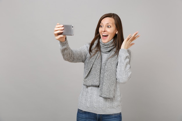 Mulher jovem animada no suéter, lenço estendendo as mãos, fazendo selfie filmado no celular, fazendo videochamada isolada no fundo cinza. conceito de estação fria de emoções de pessoas de estilo de vida de moda saudável.