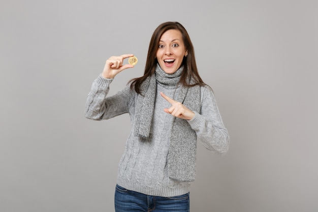 Mulher jovem animada num suéter cinza, lenço apontando o dedo indicador em bitcoin, moeda futura isolada no fundo da parede cinza. Estilo de vida de moda saudável, emoções sinceras de pessoas, conceito de estação fria.