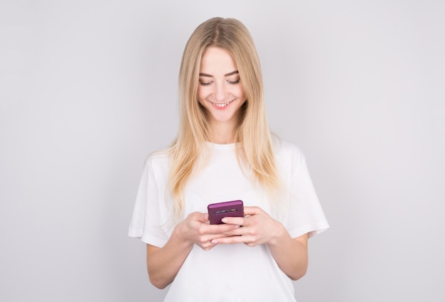 Mulher jovem animada olhando para o celular dela sorrindo. Mulher lendo mensagem de texto em seu telefone, isolada sobre uma parede branca.