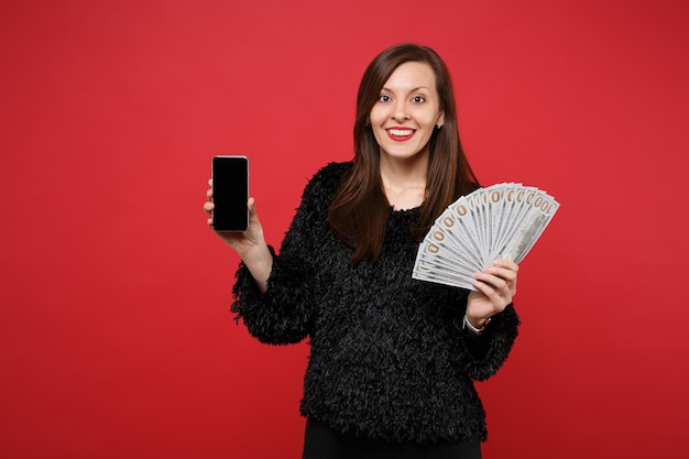 Mulher jovem animada segurando o telefone móvel com o fã de tela vazia em branco de dinheiro em notas de dólar dinheiro em dinheiro isolado sobre fundo vermelho. Emoções sinceras de pessoas, conceito de estilo de vida. Simule o espaço da cópia.