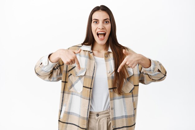 Foto mulher jovem animada, sorrindo e apontando para baixo com uma cara surpresa e feliz, mostrando uma nova oferta promocional incrível, aponte para o banner do logotipo com uma grande oferta, em pé sobre uma parede branca