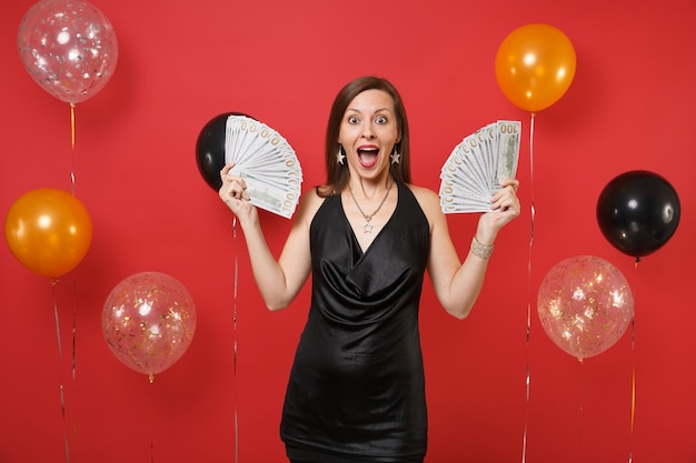 Mulher jovem animada surpresa em um vestido preto comemorando, segurando o pacote de muitos dólares, dinheiro em balões de ar de fundo vermelho brilhante. Feliz ano novo aniversário maquete feriado festa conceito.