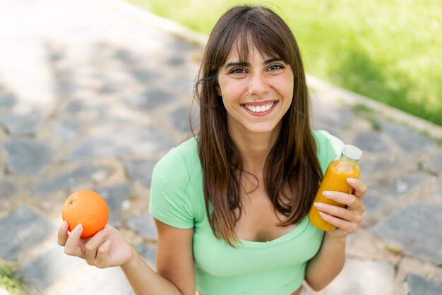 Mulher jovem ao ar livre segurando uma laranja e um suco de laranja