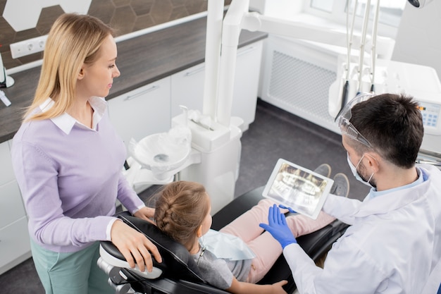 Mulher jovem ao lado da filha, sentada na poltrona do dentista, olhando para um raio-x no touchpad