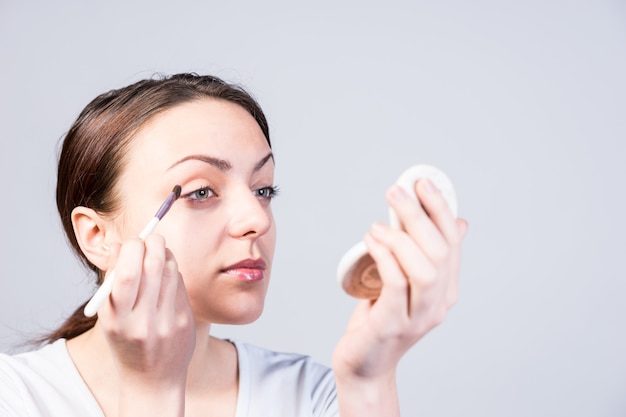Mulher jovem aplicando maquiagem nos olhos com um pó compacto na sobrancelha, sobre cinza com copyspace
