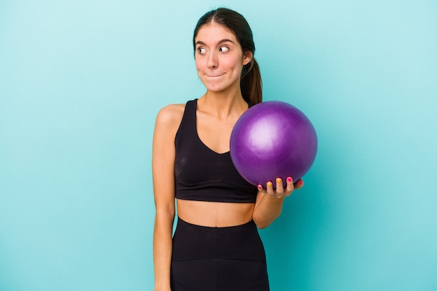 Mulher jovem aptidão caucasiana segurando uma bola isolada em um fundo azul confusa, sente-se duvidosa e insegura.