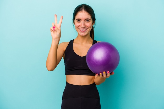 Mulher jovem aptidão caucasiana segurando uma bola isolada em um fundo azul, mostrando o número dois com os dedos.