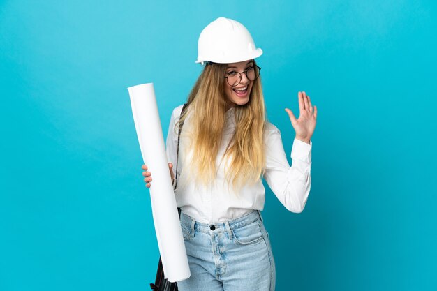 Mulher jovem arquiteta com capacete e segurando plantas isoladas na parede azul saudando com a mão com expressão feliz