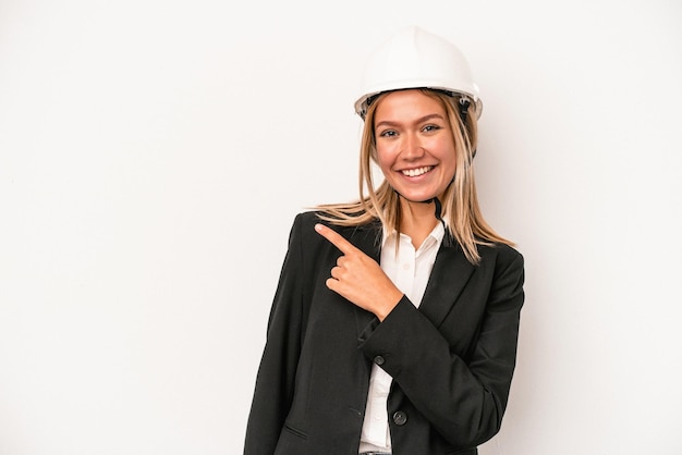 Mulher jovem arquiteto caucasiano usando um capacete isolado no fundo branco, sorrindo e apontando para o lado, mostrando algo no espaço em branco.