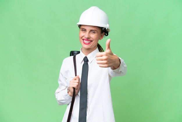 Mulher jovem arquiteto com capacete e segurando plantas sobre fundo isolado com polegares para cima porque algo de bom aconteceu