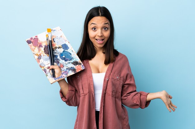 Mulher jovem artista segurando uma paleta sobre a parede azul isolada feliz e sorridente.